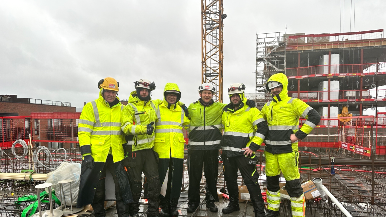 Seks arbeidere hos Østlandske VVS ser glade ut og poserer for kameraet på en byggeplass.
