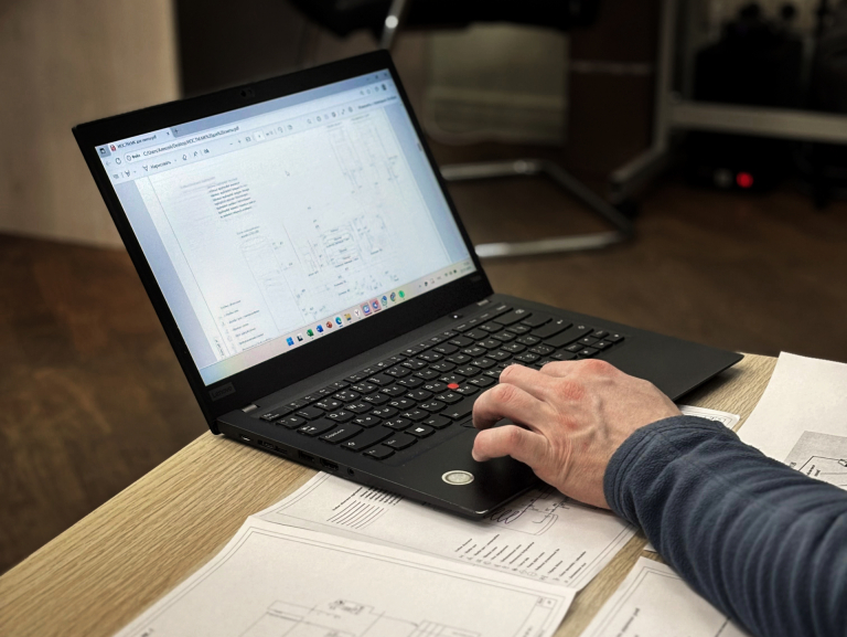 En hånd på en bærbar PC som viser en teknisk tegning, med utskrevne tegninger på bordet