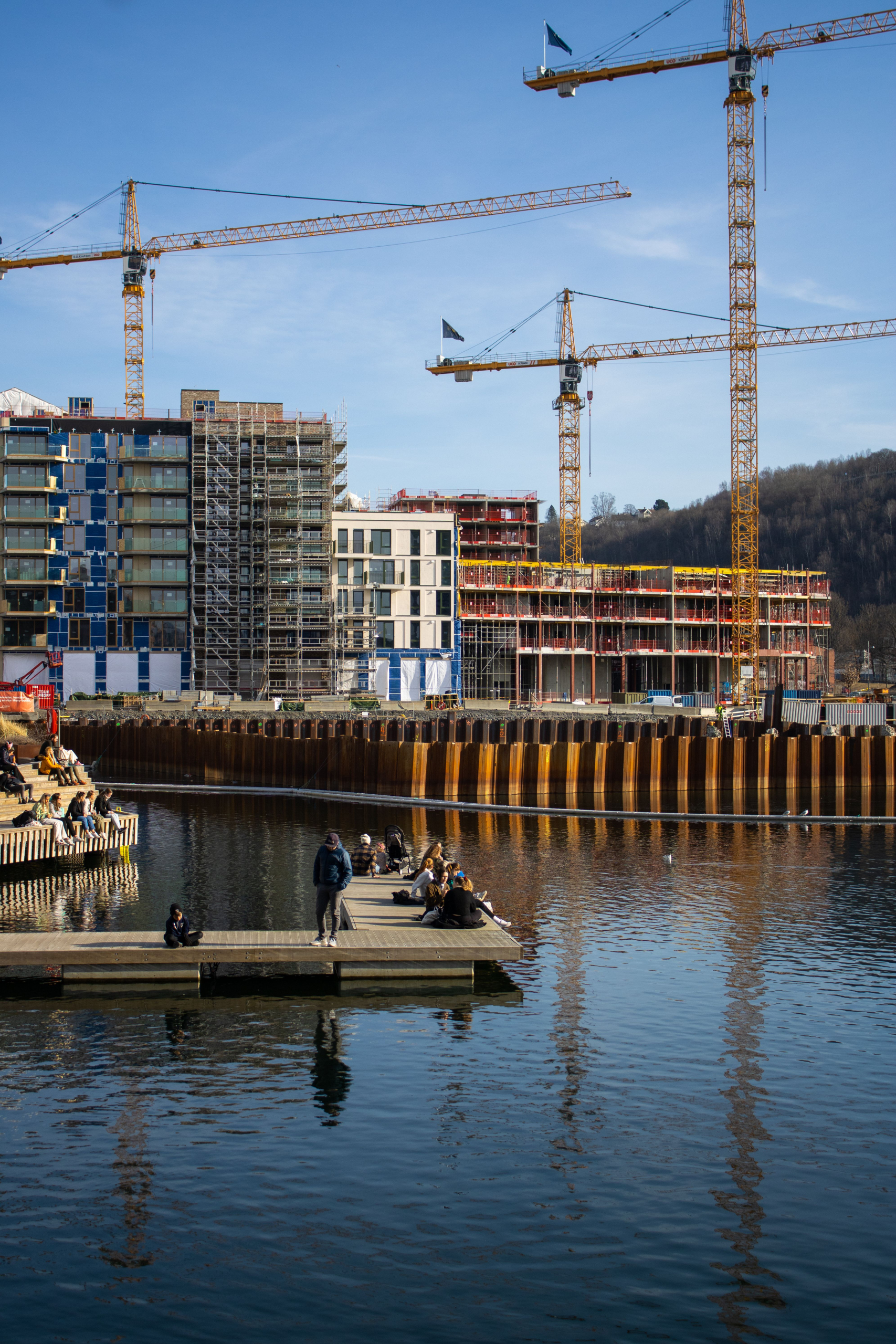 Folk på en brygge ved vannet med store byggekraner og nye boligblokker under oppføring i bakgrunnen